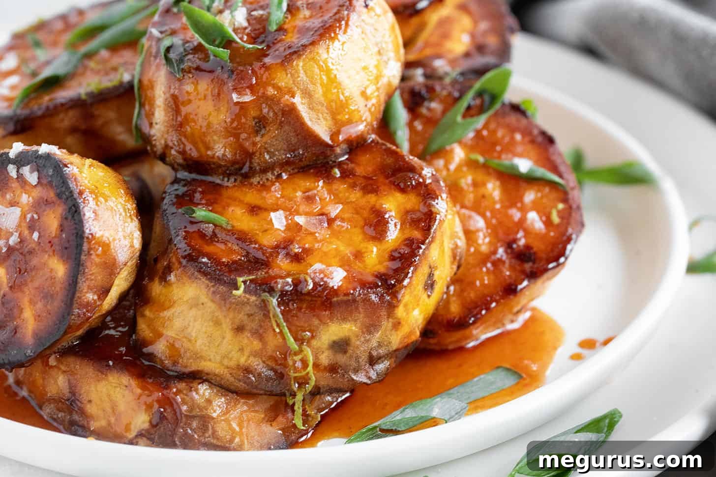 A close-up of golden-brown melting sweet potato slices drizzled with honey sriracha sauce, garnished with fresh green onions on a white plate, showcasing their crispy edges and soft interior.