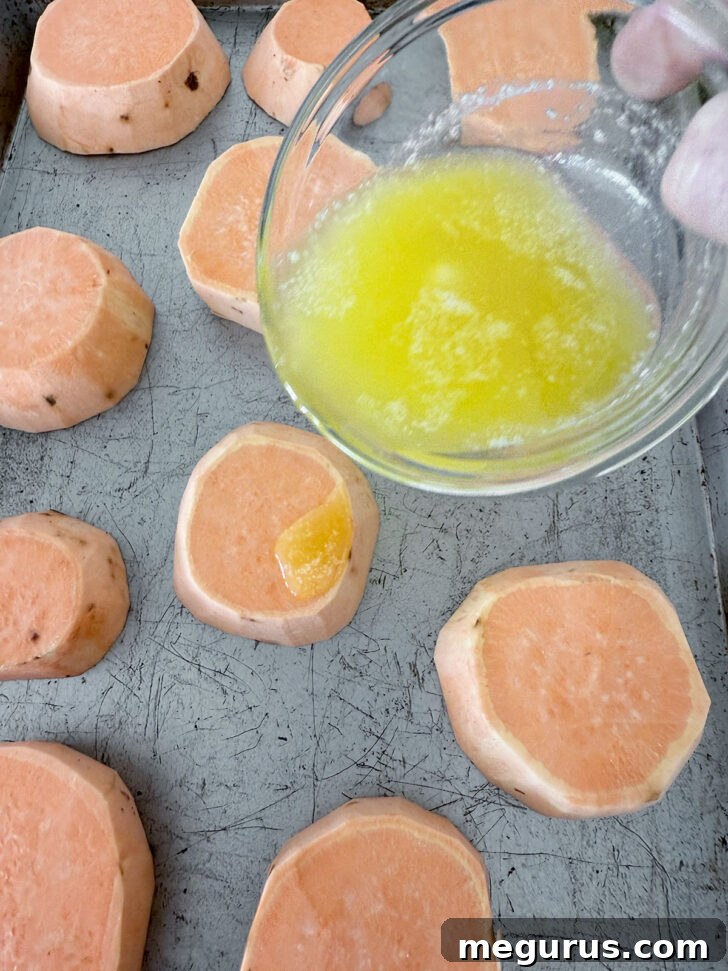 Melted butter being generously drizzled over neatly arranged sweet potato slices on a baking sheet, ensuring even coating.