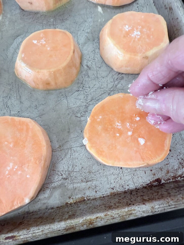 Coarse salt being sprinkled over butter-coated sweet potato slices on a baking sheet, ready for the oven.