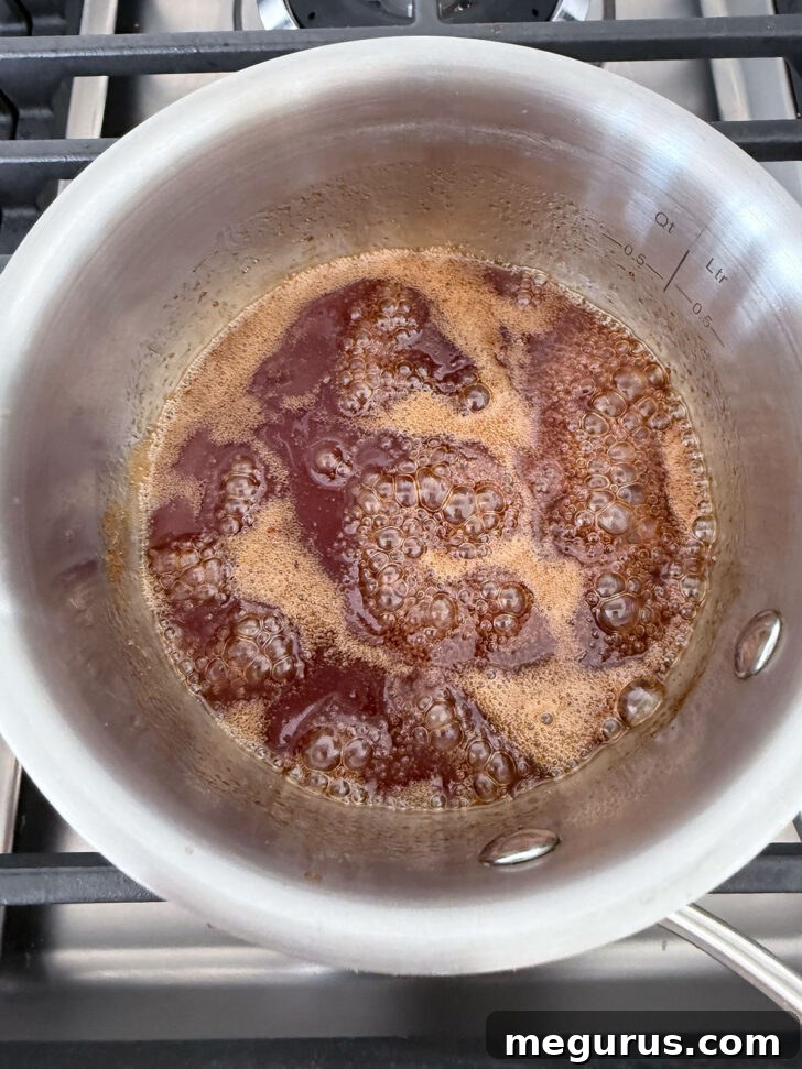 The honey sriracha sauce is simmering gently in a small saucepan on the stove, having thickened slightly after boiling.