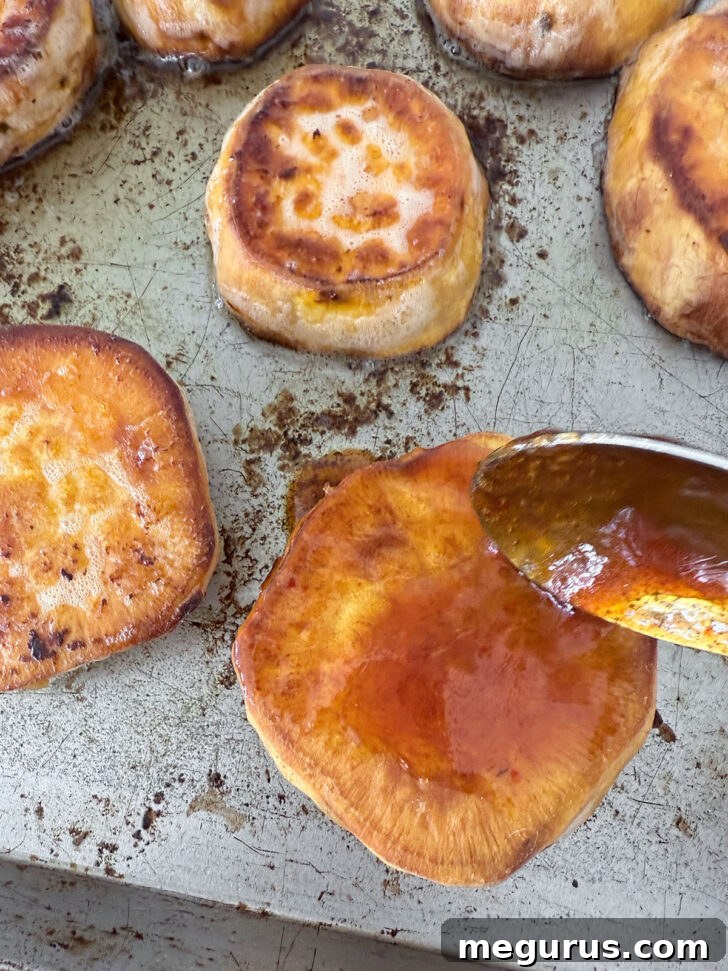 A spoon applying a small amount of the glossy honey sriracha sauce onto the roasting sweet potato slices during the final minutes of cooking.