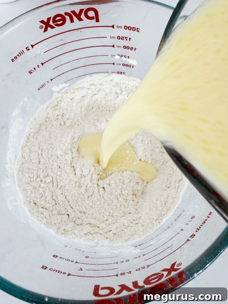 A stream of wet ingredients (eggs, oil, milk) being poured into a bowl containing dry waffle batter ingredients (flour, baking powder, salt, sugar).