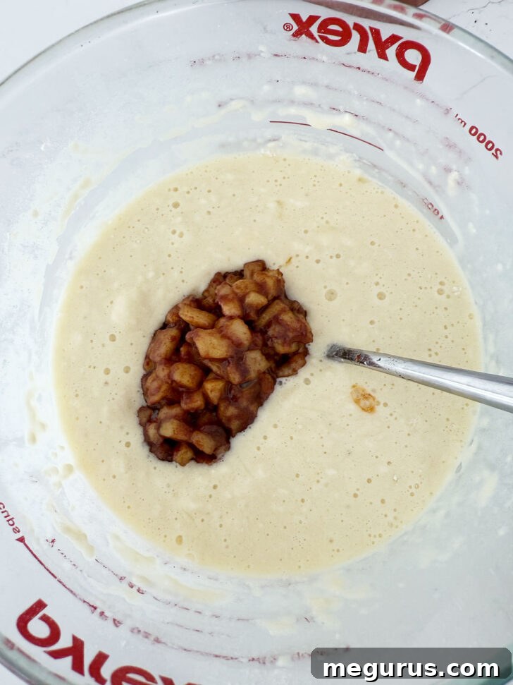 Completely cooled, cooked cinnamon apples being carefully added to the prepared waffle batter in a large mixing bowl.