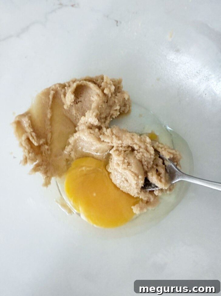 A large egg being cracked and added to the creamy pudding batter in a mixing bowl.