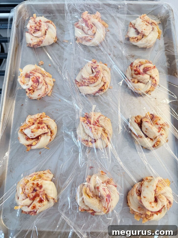 Formed pizza knots arranged on a baking sheet, covered with plastic wrap for their final rise.