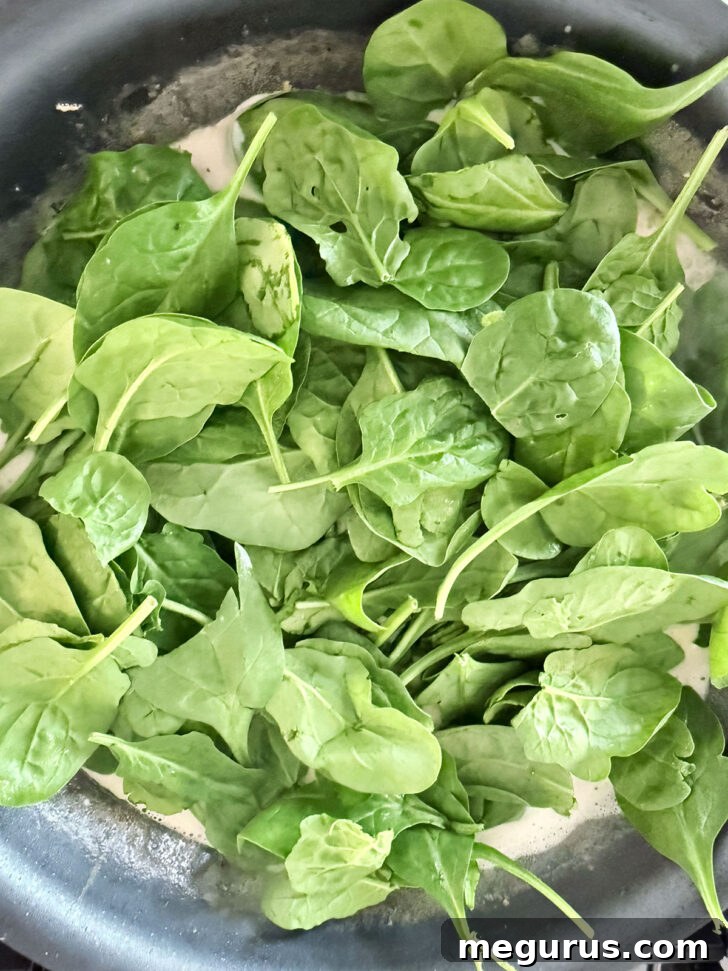 Adding fresh baby spinach to the creamy sauce in the skillet.