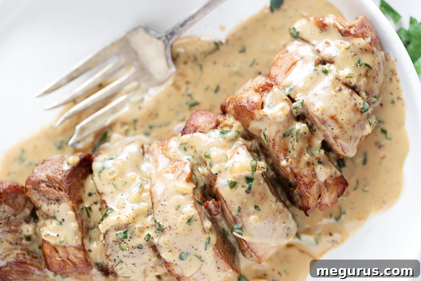 Perfectly seared pork tenderloin medallions drizzled with rich creamy Dijon sauce, garnished with fresh parsley on a white plate, ready to be served.