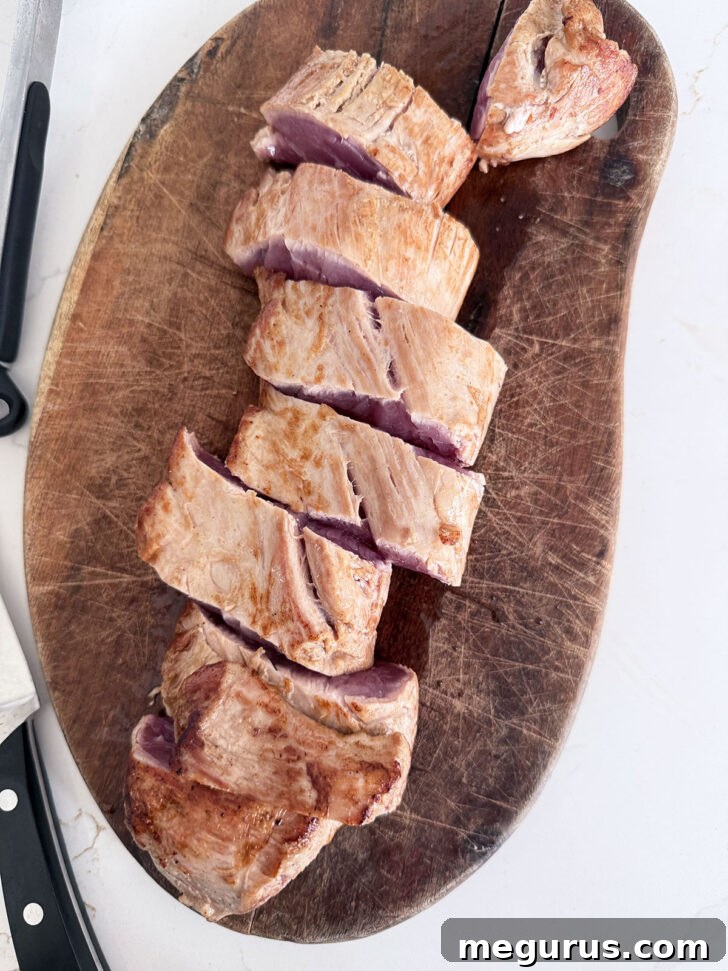 Pork tenderloin, previously seared, being sliced into even 1-inch medallions on a wooden cutting board.