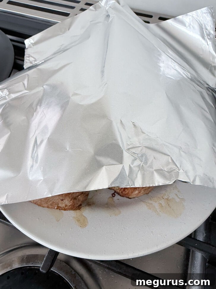 Cooked pork medallions resting on a plate, loosely covered with aluminum foil to keep warm.