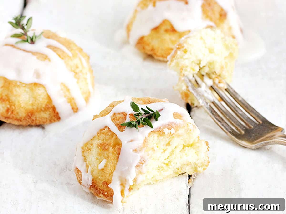 Close-up of a glazed mini lemon thyme olive oil cake on a white background, garnished with a fresh thyme sprig.