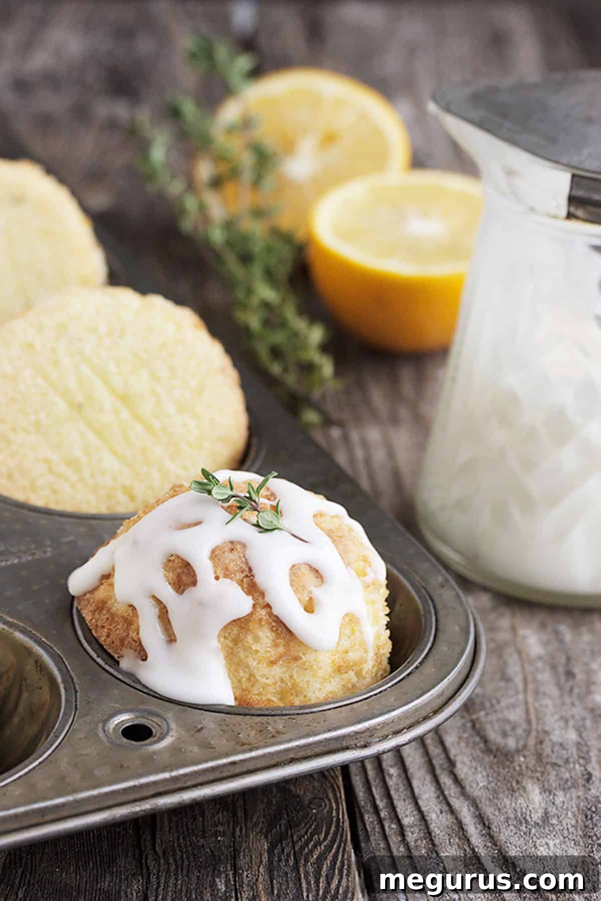 Two glazed mini lemon thyme olive oil cakes on a white background, with thyme sprigs and lemon slices.