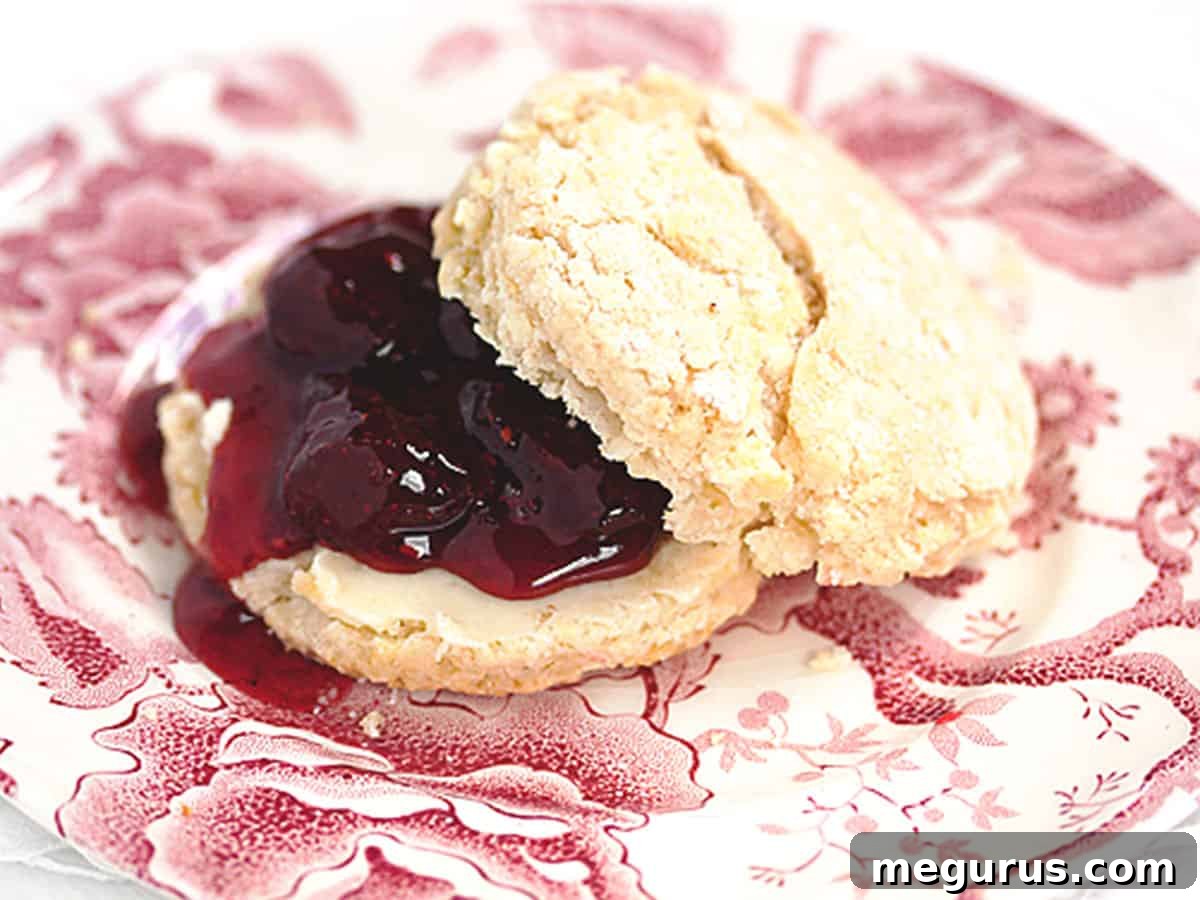 Close-up photograph of homemade strawberry jam generously spread on a warm scone, emphasizing the delicious texture and visible fruit pieces.