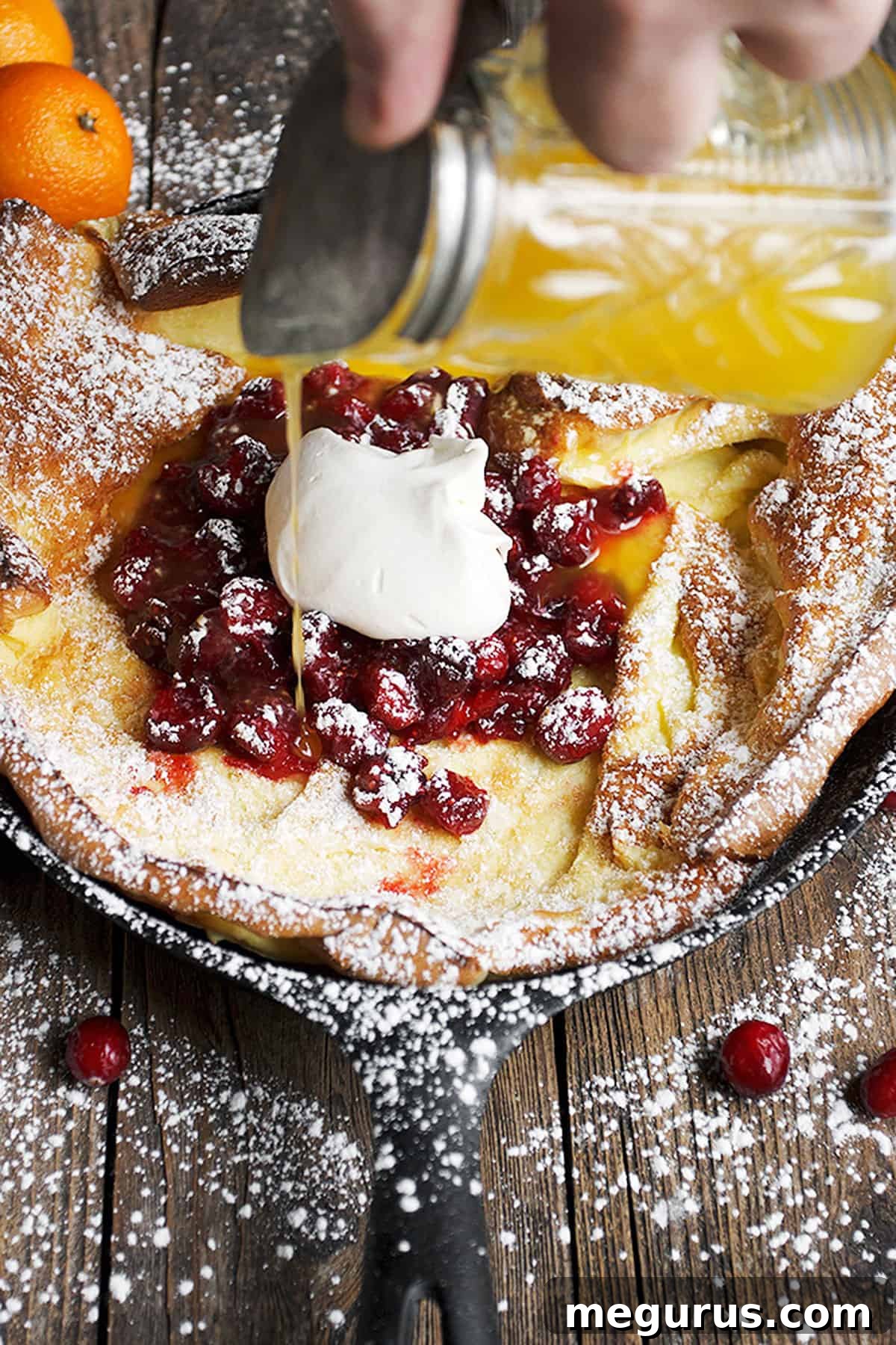 Close-up of a Cranberry Dutch Baby pancake being drizzled with clementine orange syrup, highlighting its golden crust and fruit topping.