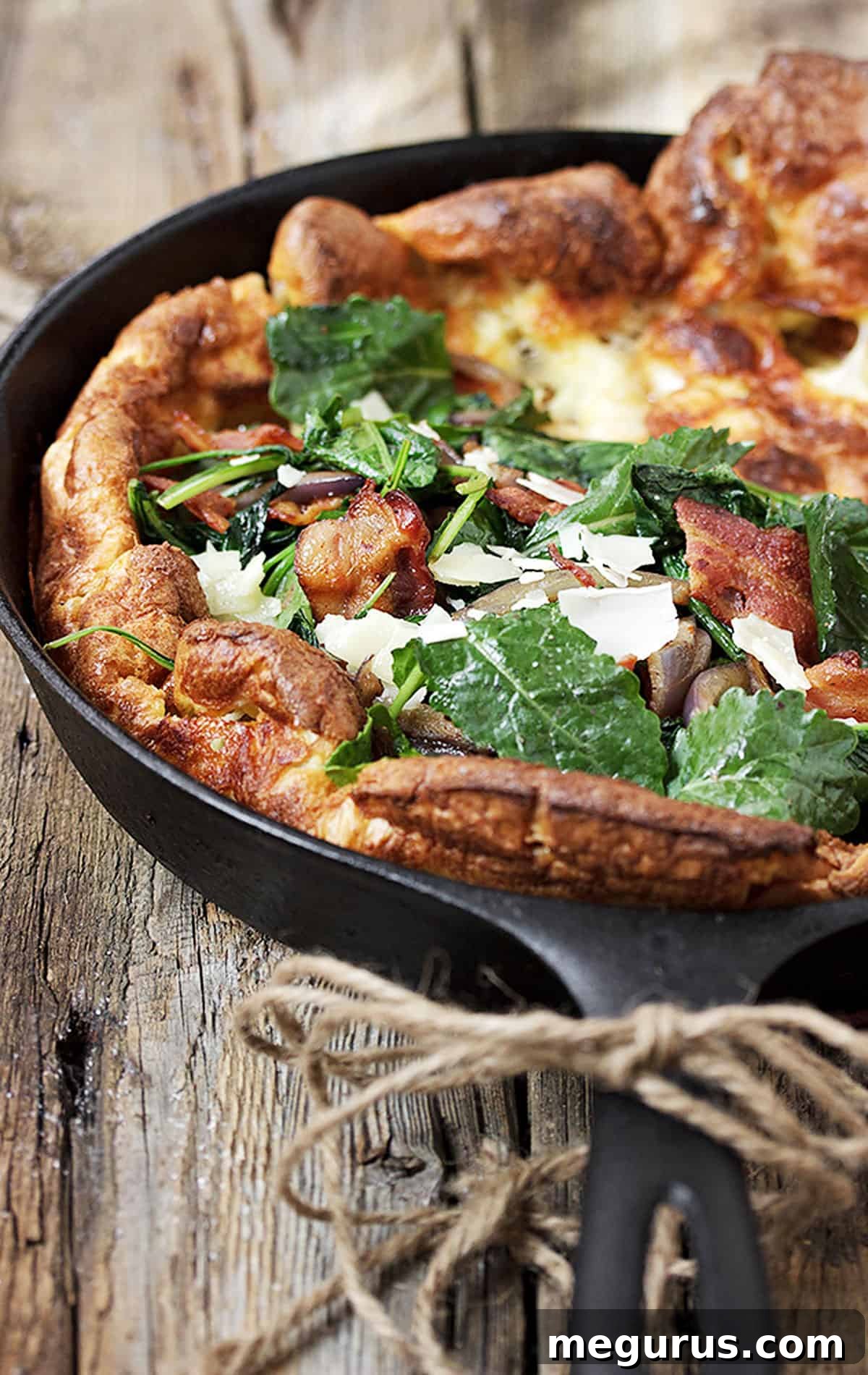 Close-up of a Kale Bacon Dutch Baby in a cast iron skillet, garnished with shaved cheddar.