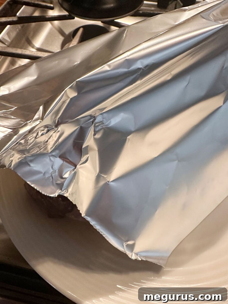The cooked pork tenderloin resting on a plate, covered loosely with aluminum foil