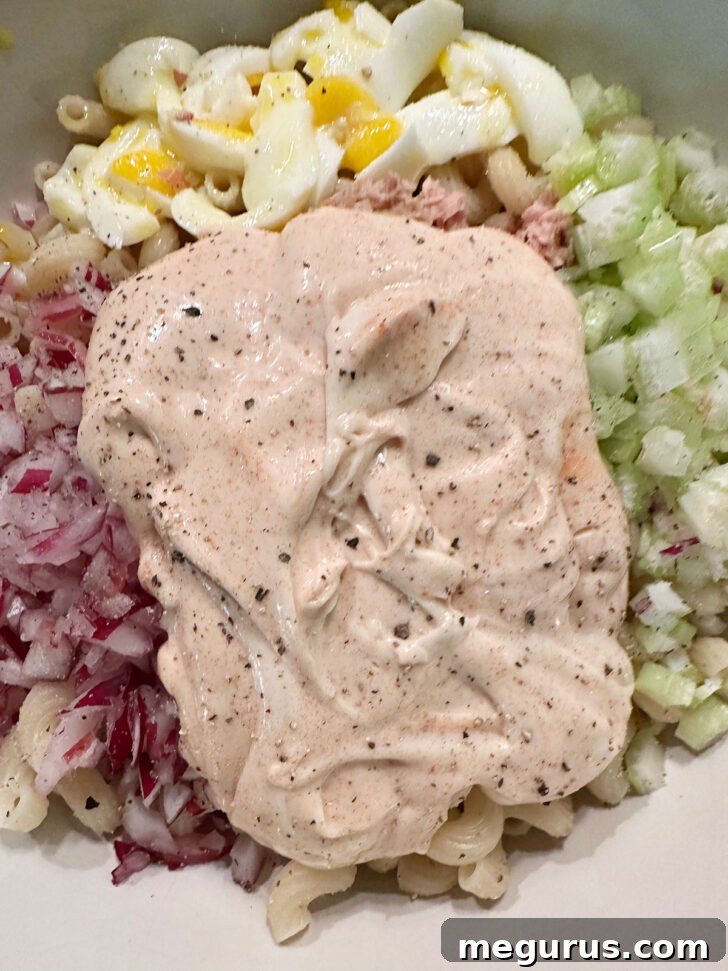 Creamy salad dressing being poured over the pasta and other ingredients in the mixing bowl.