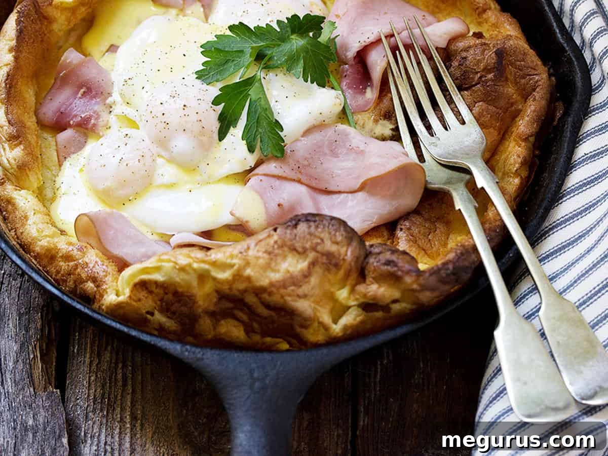 A golden, puffy Eggs Benedict Dutch Baby in a cast iron skillet, topped with ham and poached eggs, ready to be drizzled with Hollandaise sauce.