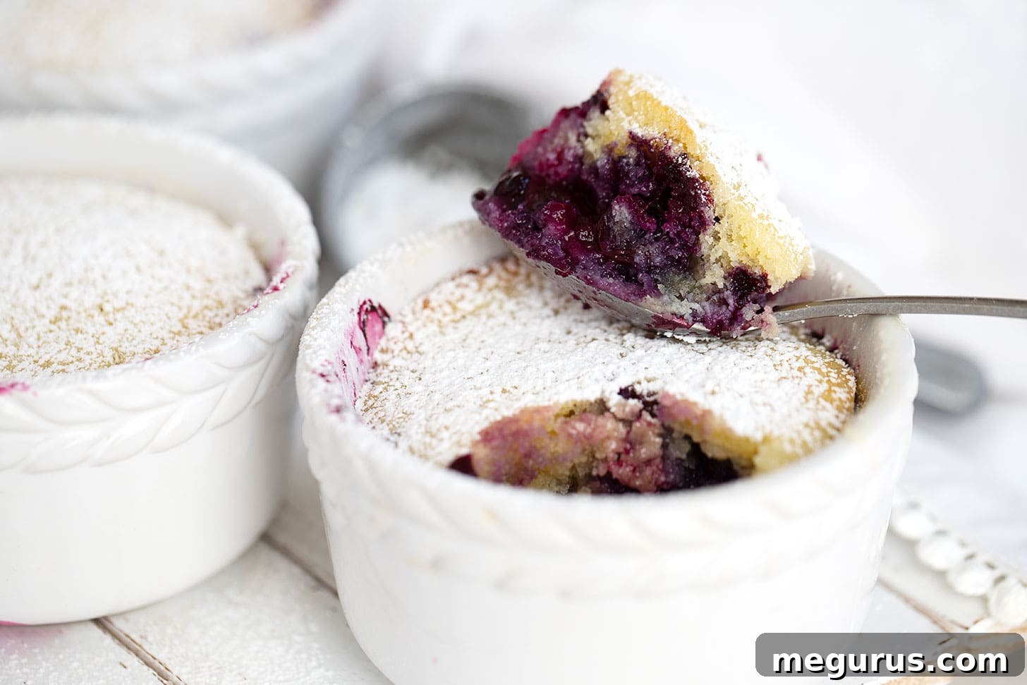 Individual blueberry pudding cakes in ramekins, served with a spoon, highlighting the warm, saucy blueberry layer beneath the golden cake.