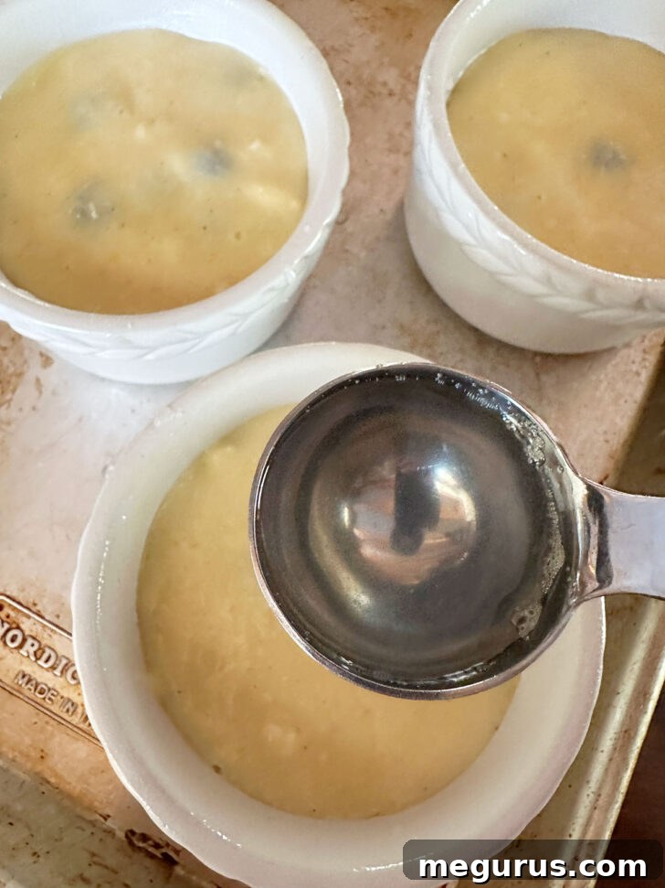 Hot lemon sugar syrup being carefully spooned over the golden cake batter, which covers a layer of blueberries in a ramekin.