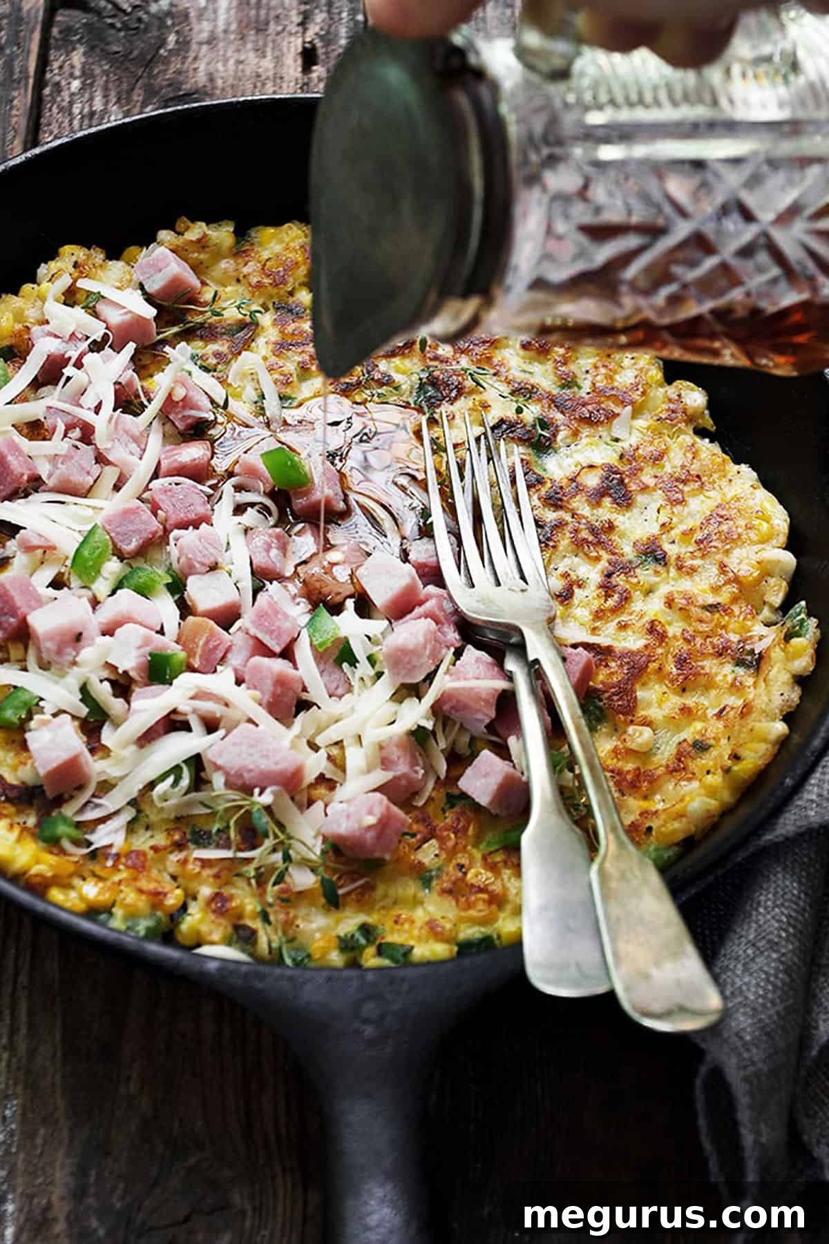 Savoury corn pancake in a cast iron skillet, generously topped with delicious cubed ham and rich, melted aged cheddar cheese, inviting a taste of a hearty brunch.