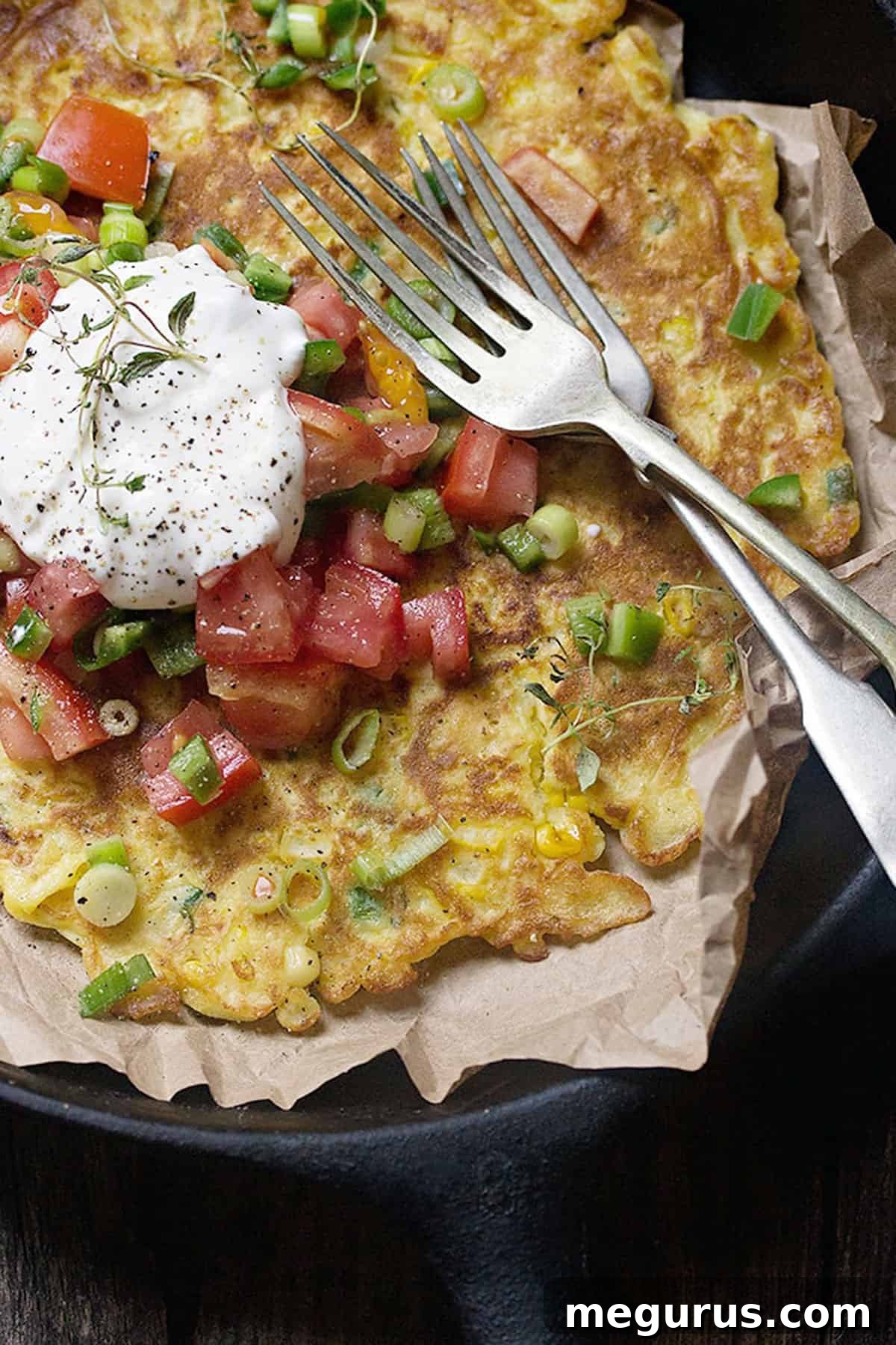 A freshly cooked savoury corn pancake in a cast iron skillet, expertly garnished with a vibrant, colorful fresh salsa featuring diced tomatoes, sliced green onions, and fresh jalapeños, with a dollop of sour cream on the side, ready to be enjoyed as a gourmet meal.