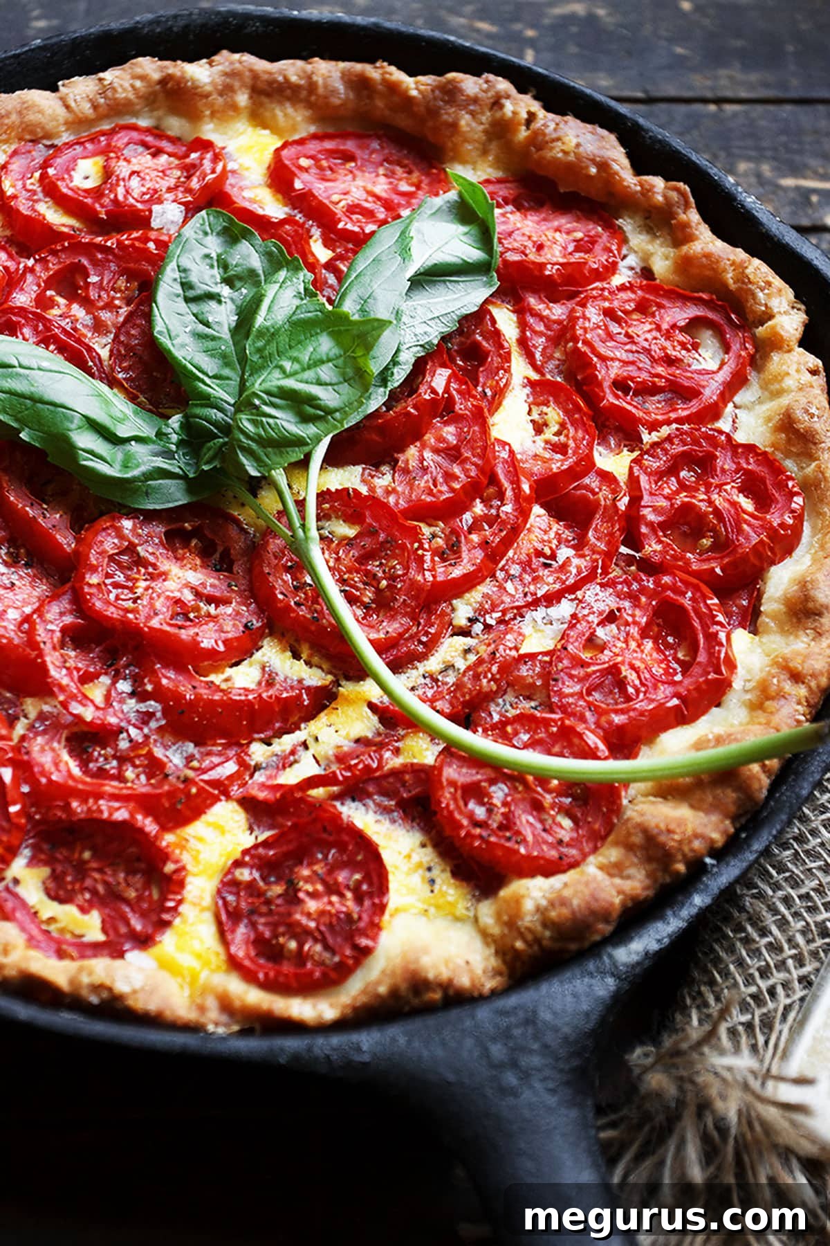 A slice of tomato pie being removed from a cast iron skillet, revealing layers of crust, cheese, basil, and tomatoes.