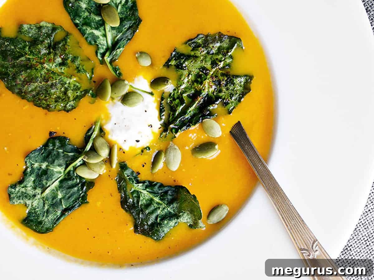 Vibrant roasted carrot ginger soup, beautifully garnished with kale chips, whipped goat cheese, and pumpkin seeds, perfect for a cozy fall meal.