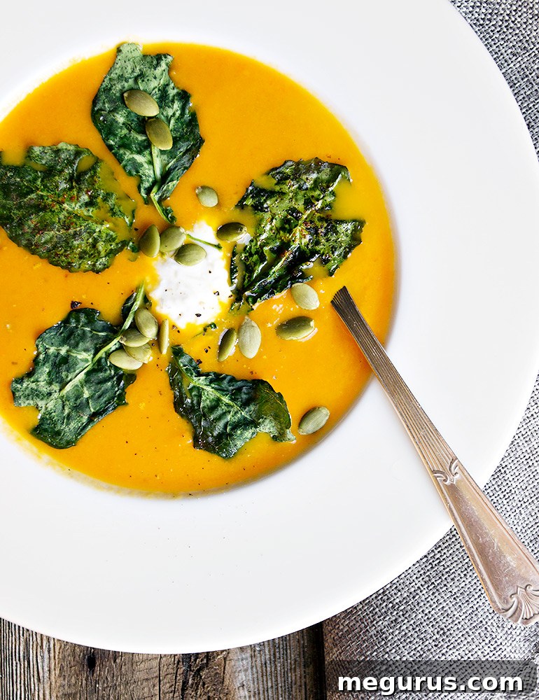 Close-up of a serving of Roasted Carrot and Ginger Soup, garnished with vibrant green kale chips and a dollop of creamy whipped goat cheese, in a rustic bowl.