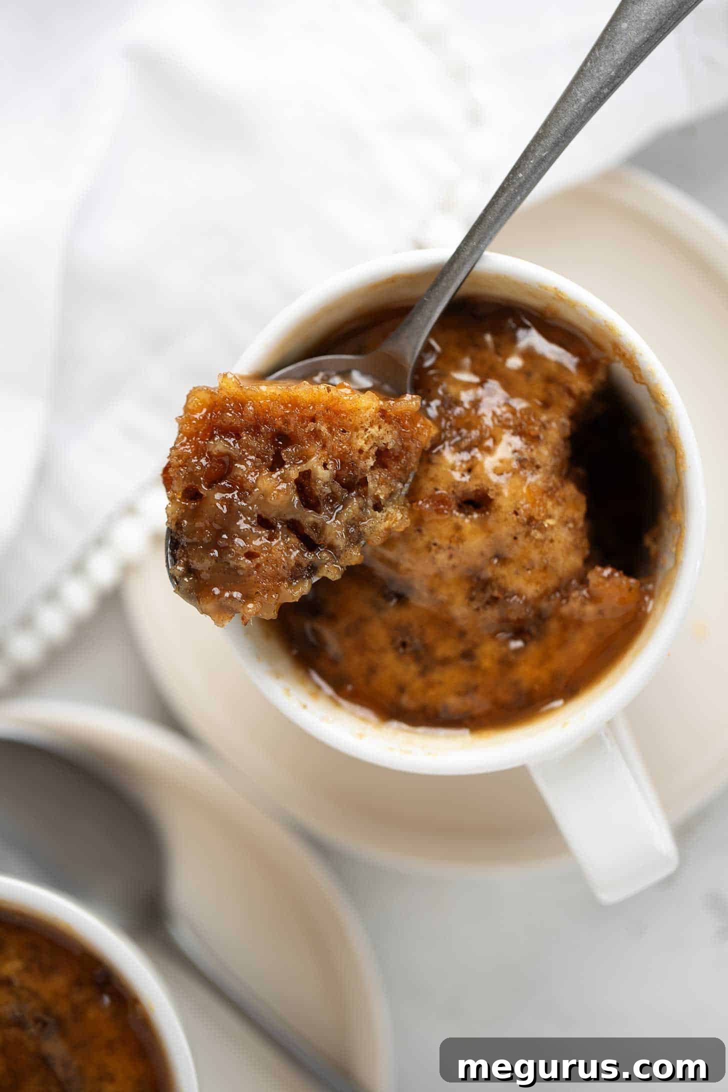 Close-up of a microwave sticky toffee pudding in a coffee cup, smothered in sticky toffee sauce, with a spoon ready to dive in