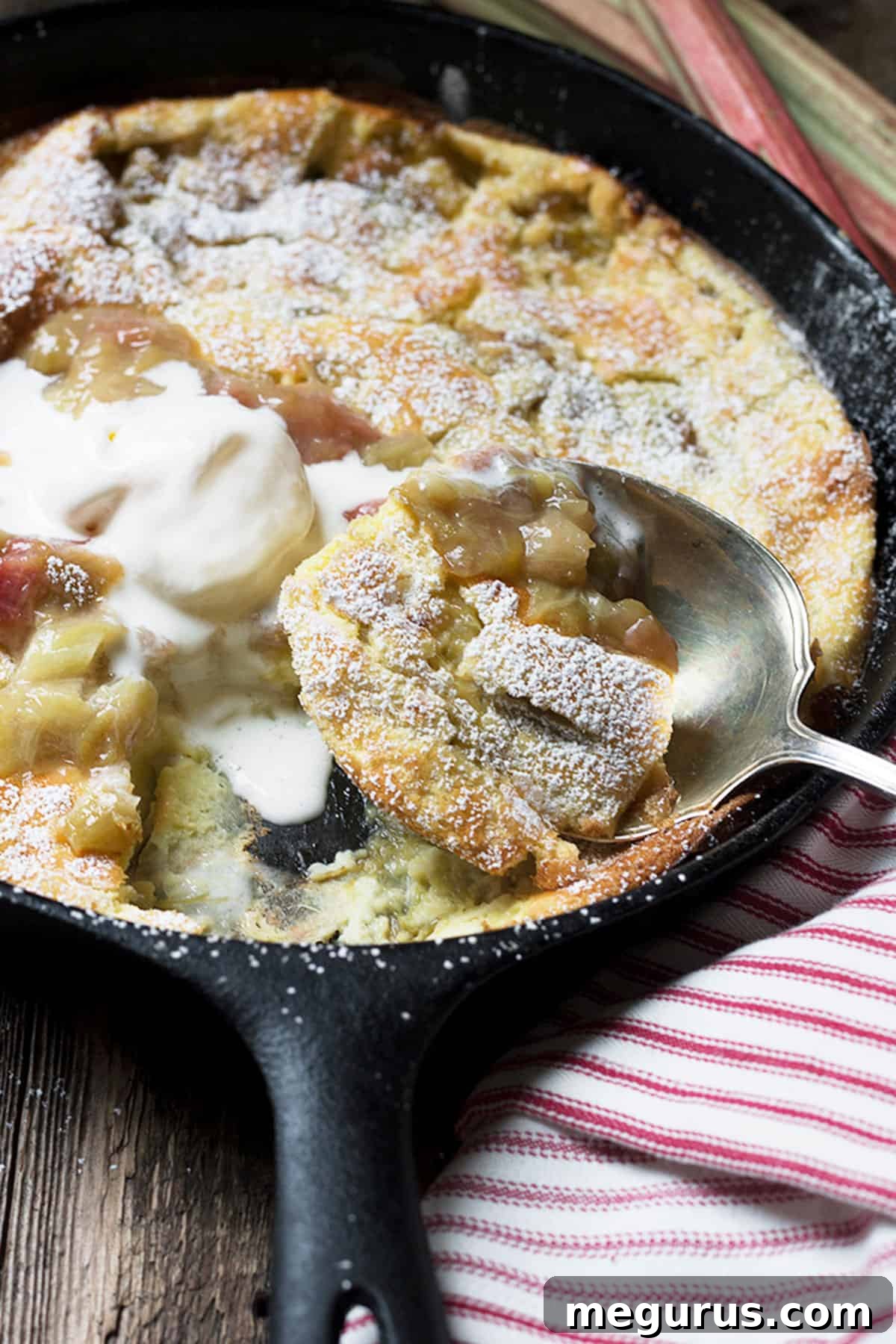 A close-up of a slice of Rhubarb Dutch Baby served in a cast iron skillet, showcasing the custardy interior and vibrant rhubarb topping.