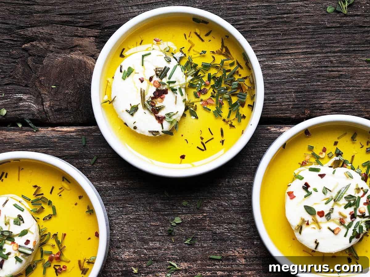 Marinated goat cheese in small white bowls with a serving knife, surrounded by fresh herbs