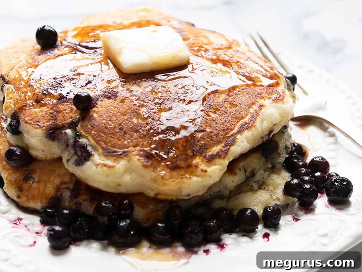 Stack of fluffy buttermilk blueberry pancakes on a plate, garnished with fresh blueberries and syrup.