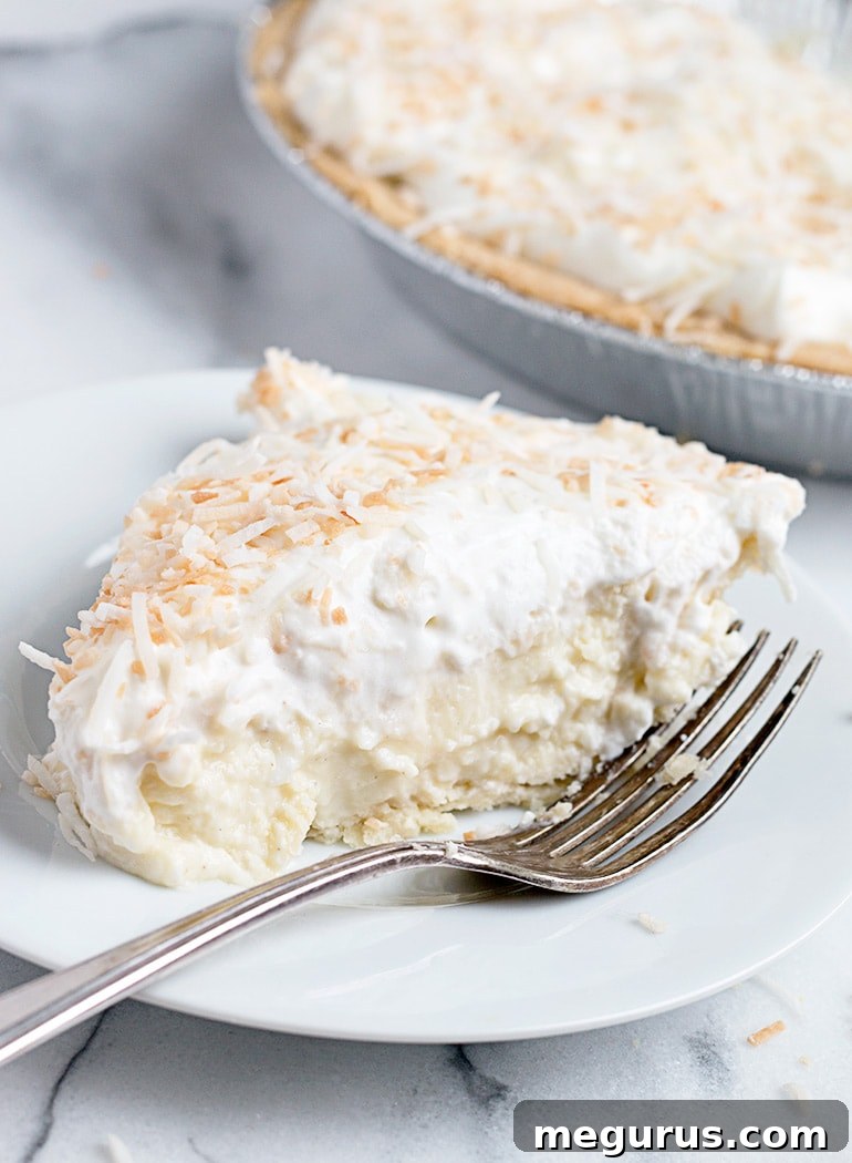 A beautifully sliced homemade coconut cream pie on a white plate, revealing its creamy layers and golden crust, topped with light whipped cream and toasted coconut.