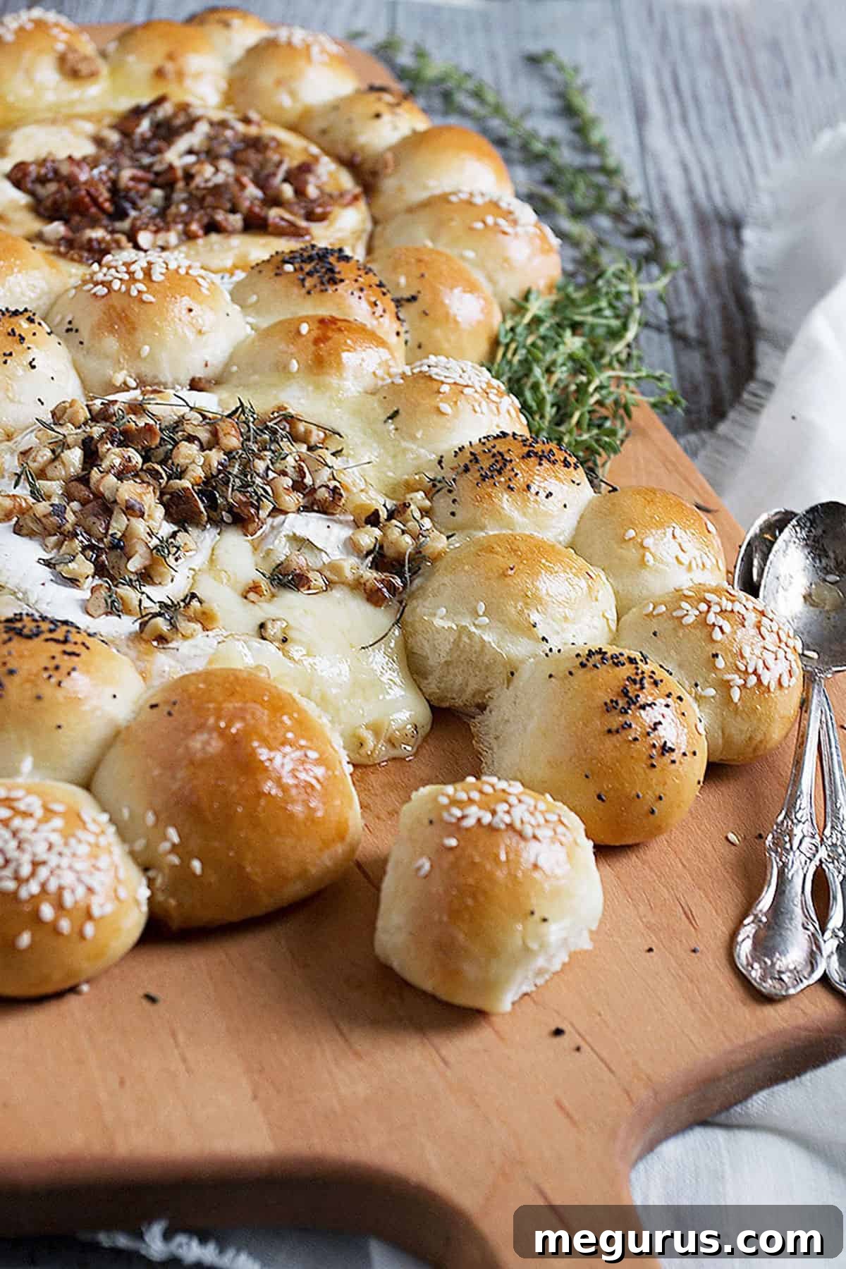 A close-up view of a baked Brie and Camembert appetizer surrounded by warm, golden bread rolls, garnished with herbs and nuts, presented on a rustic wooden board.