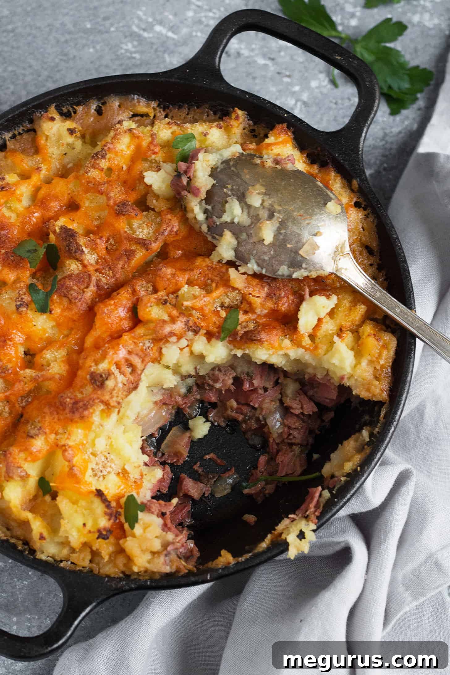 A close-up shot of a golden-brown Corned Beef Cottage Pie in a skillet, showcasing its crispy potato topping and bubbling filling, with a spoon digging in.