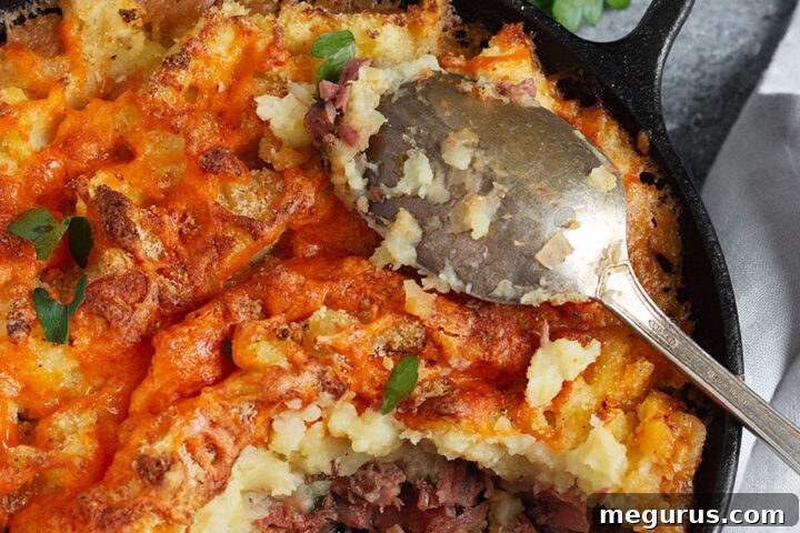 A delicious Corned Beef Cottage Pie in a skillet with a serving spoon, showcasing its golden mashed potato topping.