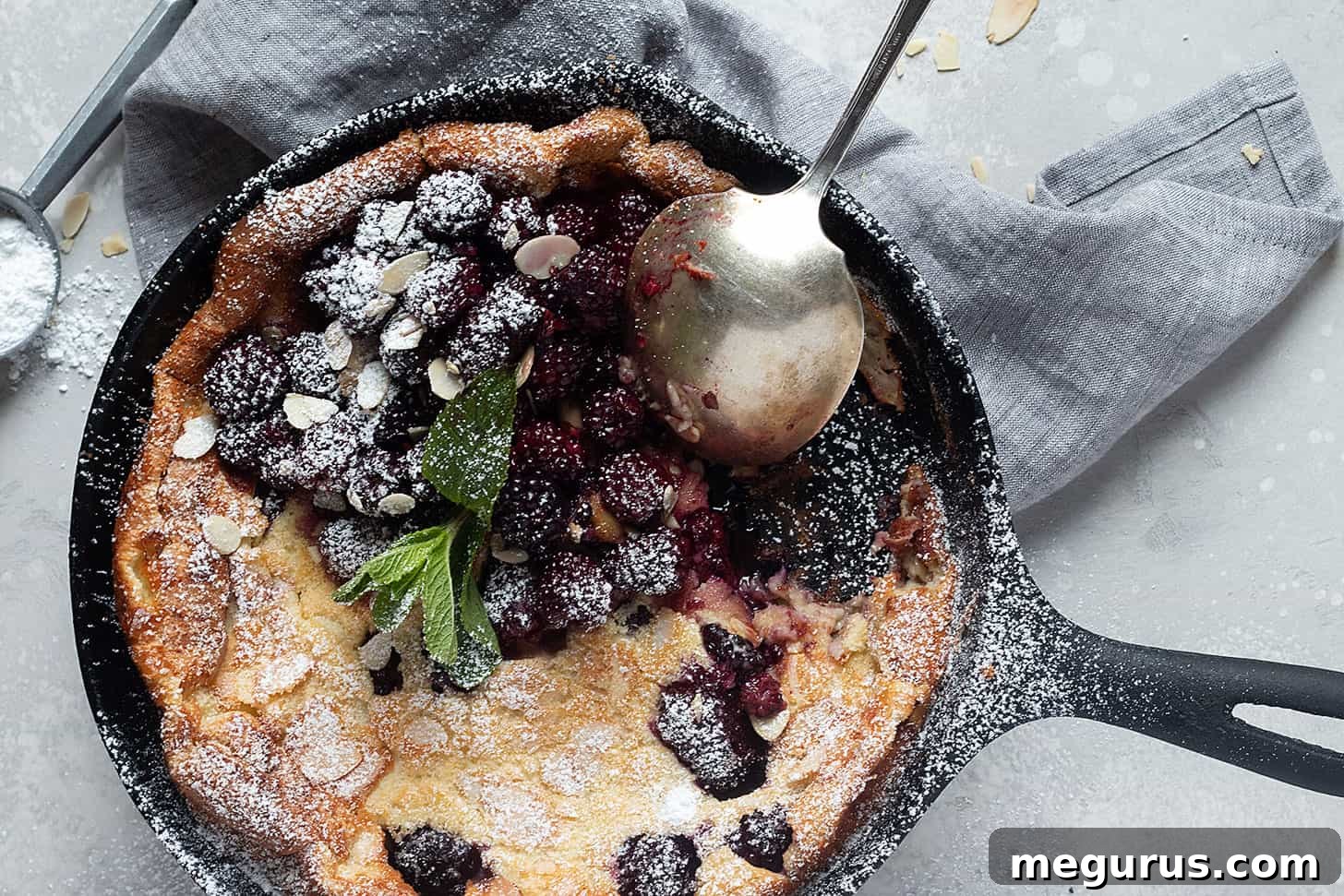 A golden-brown Blackberry Dutch Baby in a cast iron skillet, topped with roasted blackberries, sliced almonds, and a dusting of powdered sugar, with a spoon ready to serve.