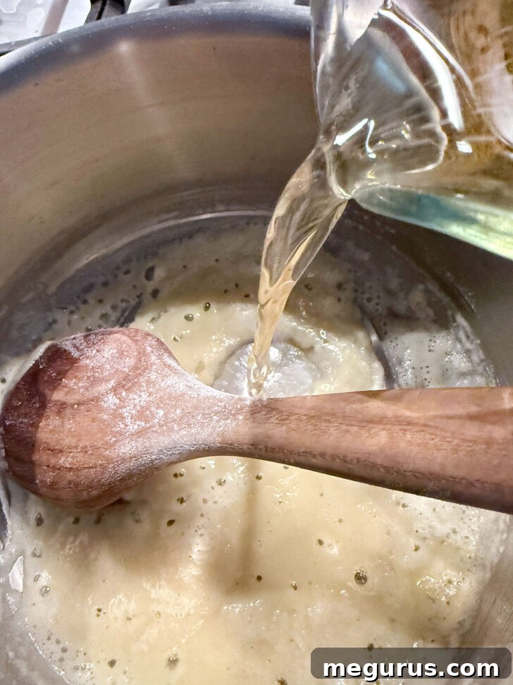 Beer being gradually poured into the roux in a saucepan, while stirring.