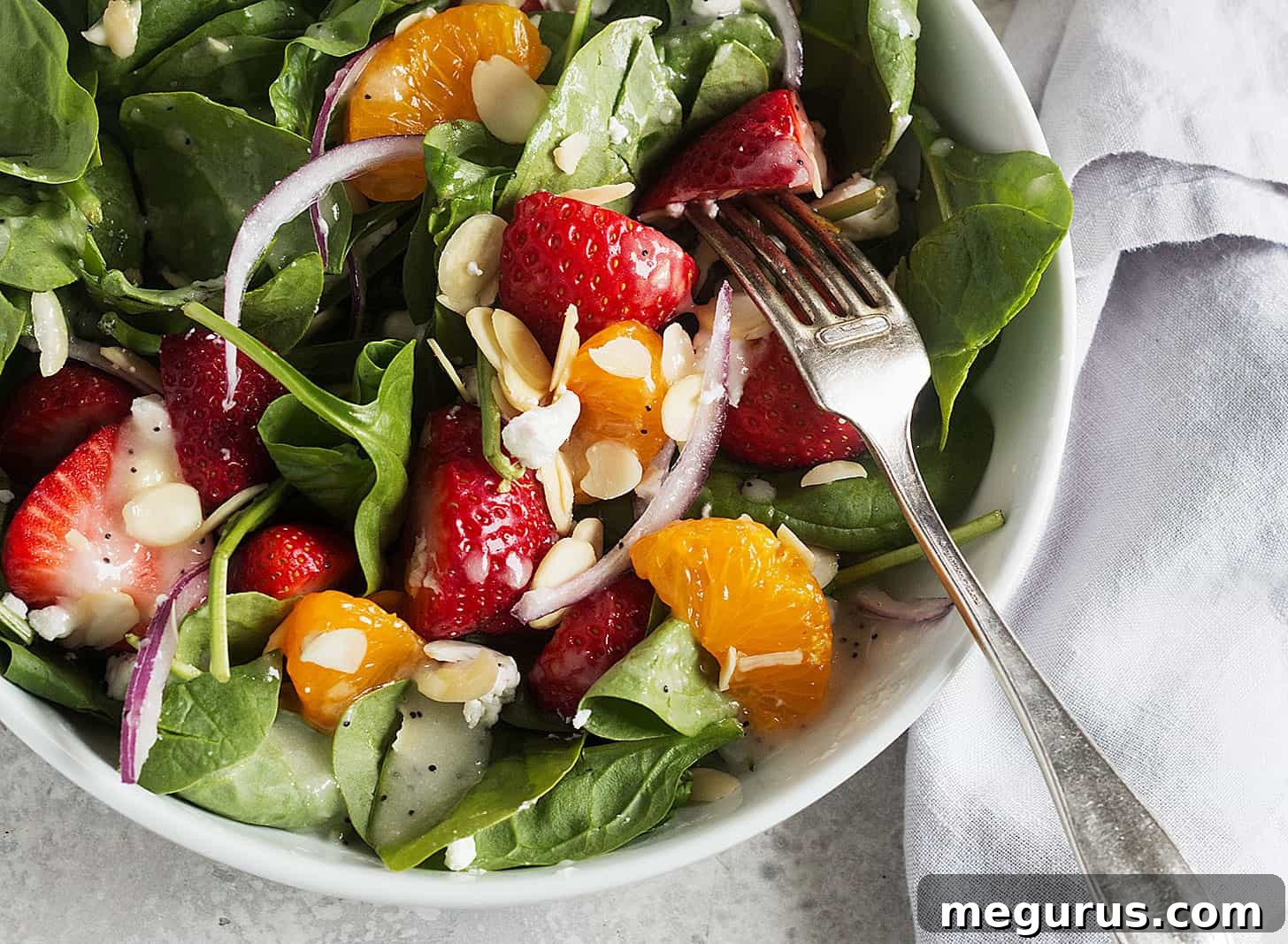 A fresh and vibrant strawberry spinach mandarin salad in a large serving bowl, with visible strawberries, mandarin segments, goat cheese, and almonds, dressed with poppy seed dressing.