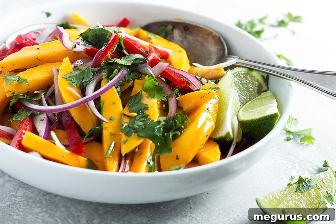 A vibrant mango salad in a serving bowl with a spoon, showcasing fresh mango slices, red bell peppers, and herbs.