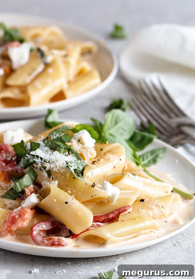 Close-up of creamy pasta adorned with roasted red peppers, cherry tomatoes, crumbled goat cheese, grated Parmesan, and vibrant fresh basil leaves.