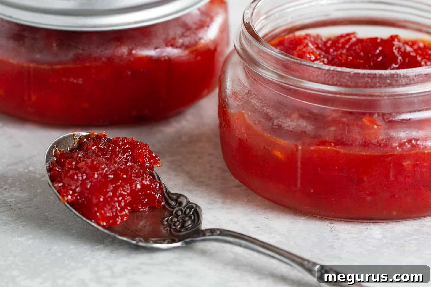 Two jars of vibrant homemade red pepper jam with a spoon full of jam, showcasing its smooth texture and rich color.