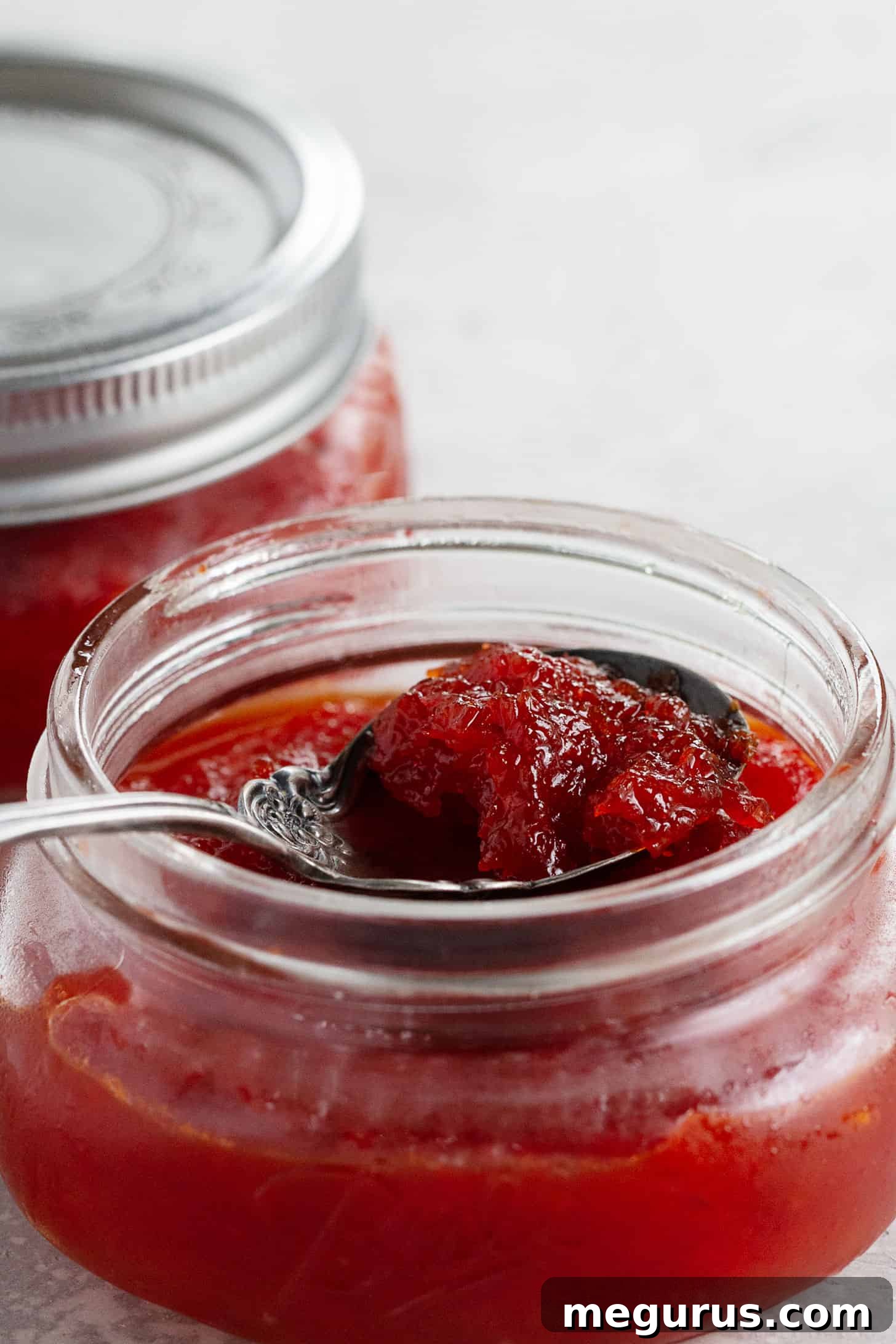 Freshly prepared red pepper jam beautifully displayed in two clear jars, with a spoon ready for tasting on a rustic surface.