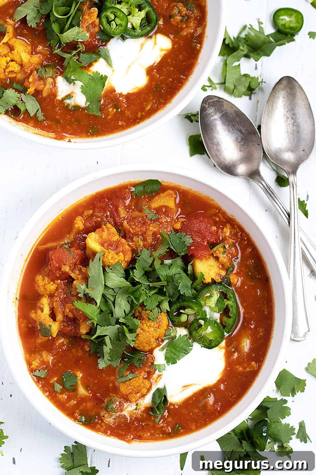 Two white bowls filled with Indian cauliflower soup, garnished with cilantro and sliced jalapenos, with spoons.
