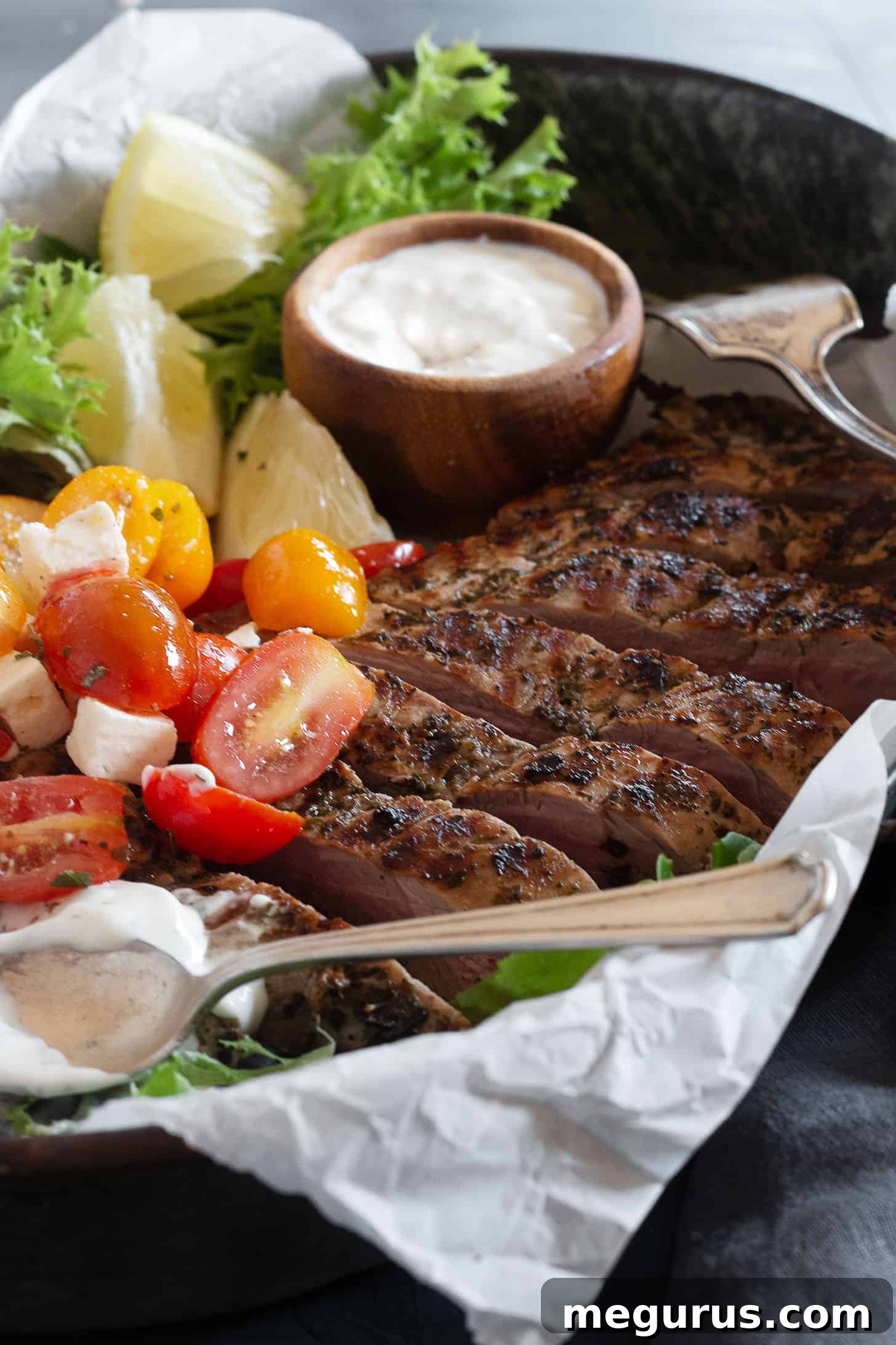 Sliced Greek Souvlaki pork tenderloin on a serving platter, garnished with fresh herbs, alongside tzatziki and a colorful tomato salad, ready to be enjoyed.