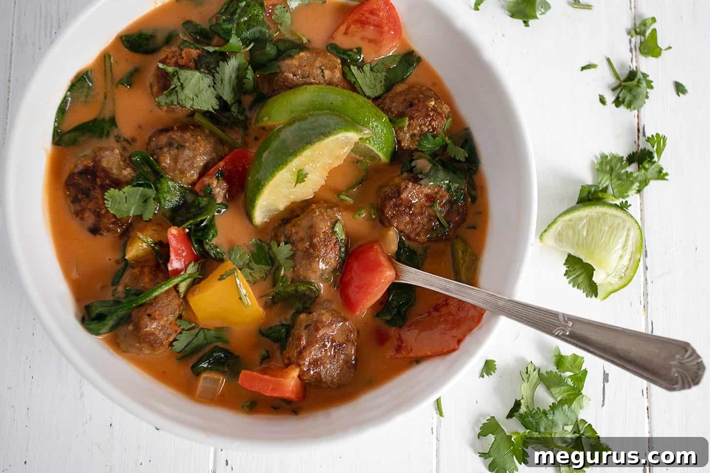 A steaming bowl of vibrant Thai chicken meatball soup, garnished with fresh cilantro and a lime wedge, with a spoon resting in it.