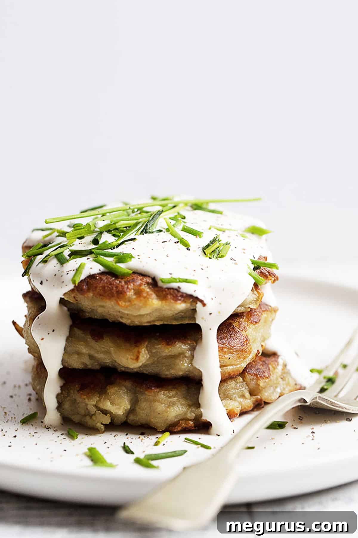 Delicious Boxty Irish potato cakes stacked neatly on a white plate, garnished with fresh sour cream and chopped green onions, ready to be enjoyed.