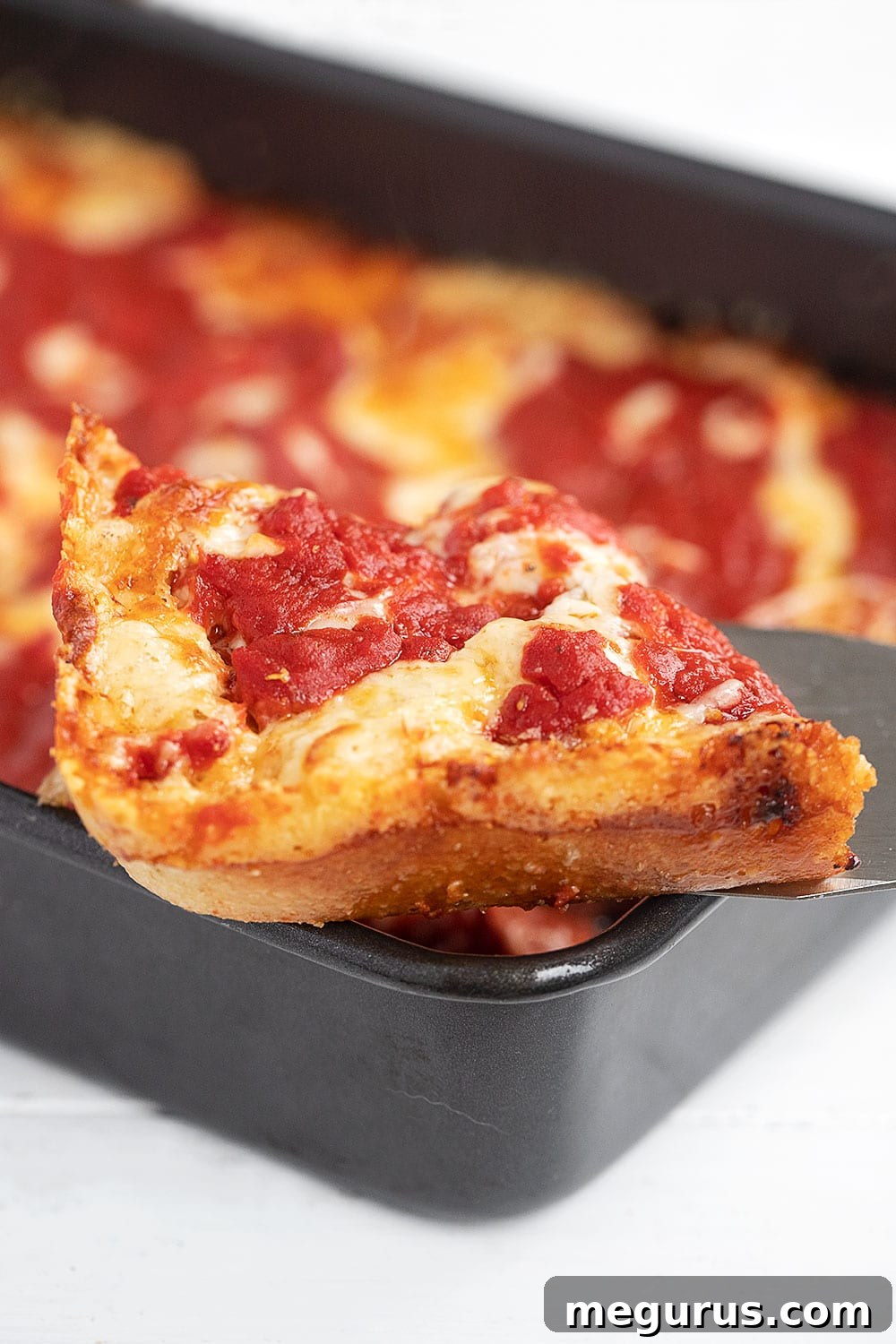 A perfectly cooked slice of Detroit Style Pizza on a spatula, showcasing the thick, airy crust and the famous crispy, caramelized cheese edges.