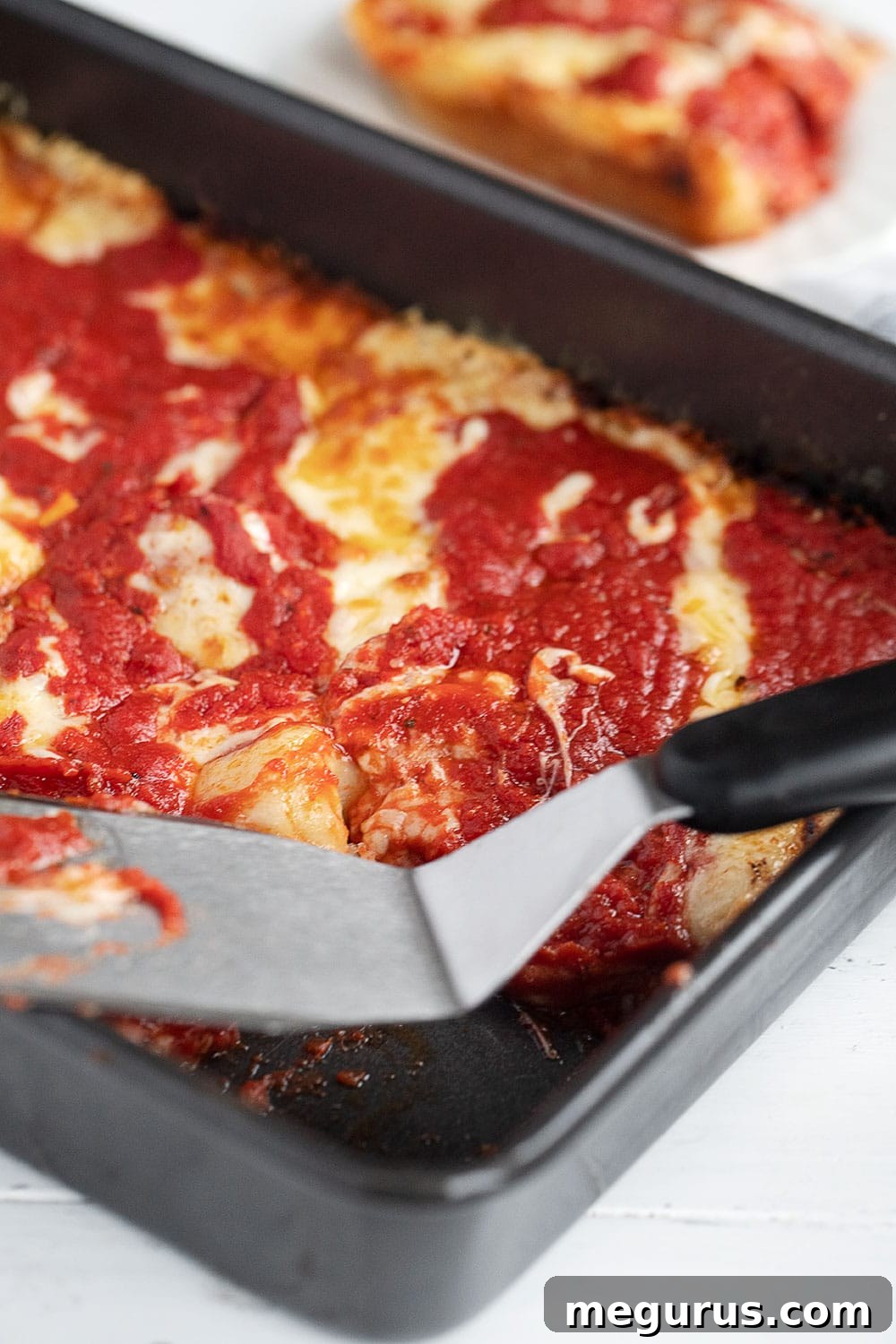 A delicious Detroit Style pizza fresh from the oven, still in its pan, with a spatula ready to serve a square slice.