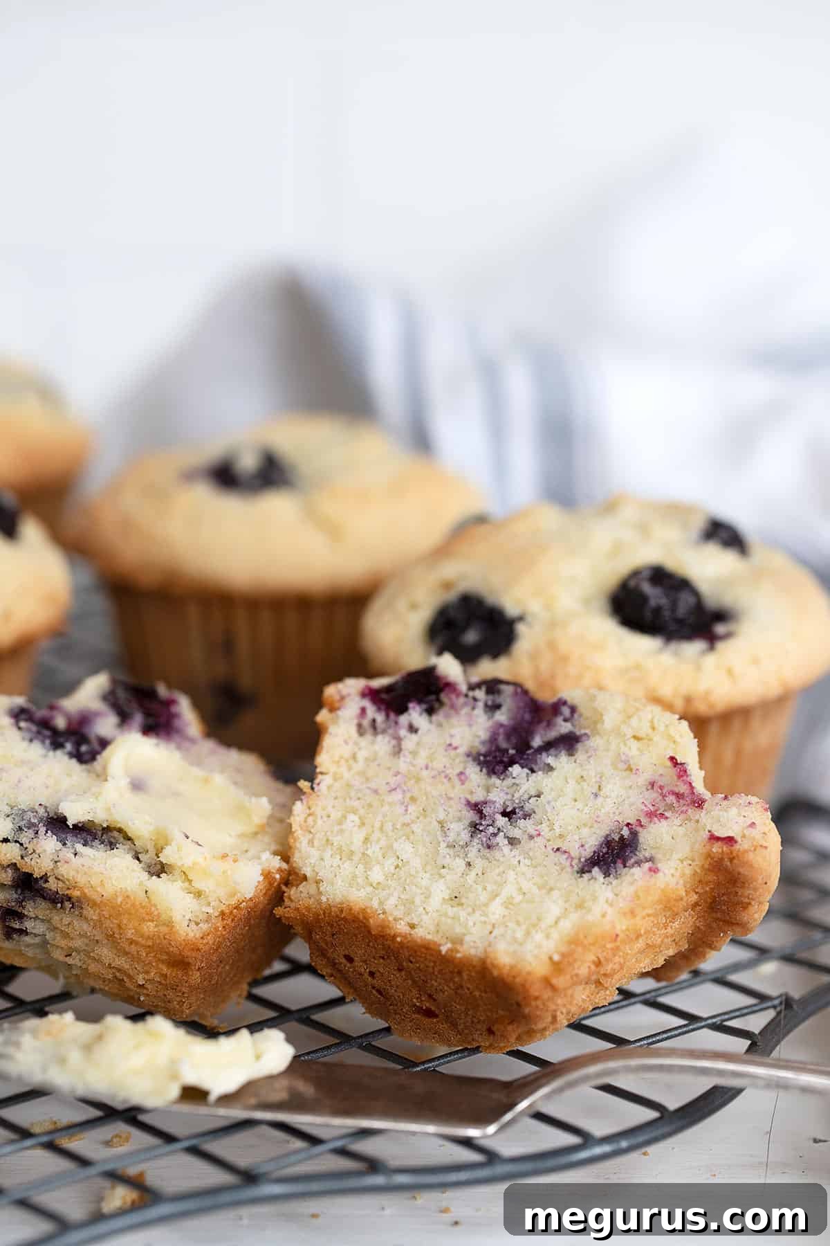 A delicious buttermilk blueberry muffin, cut open and buttered, resting on a cooling rack with other muffins.
